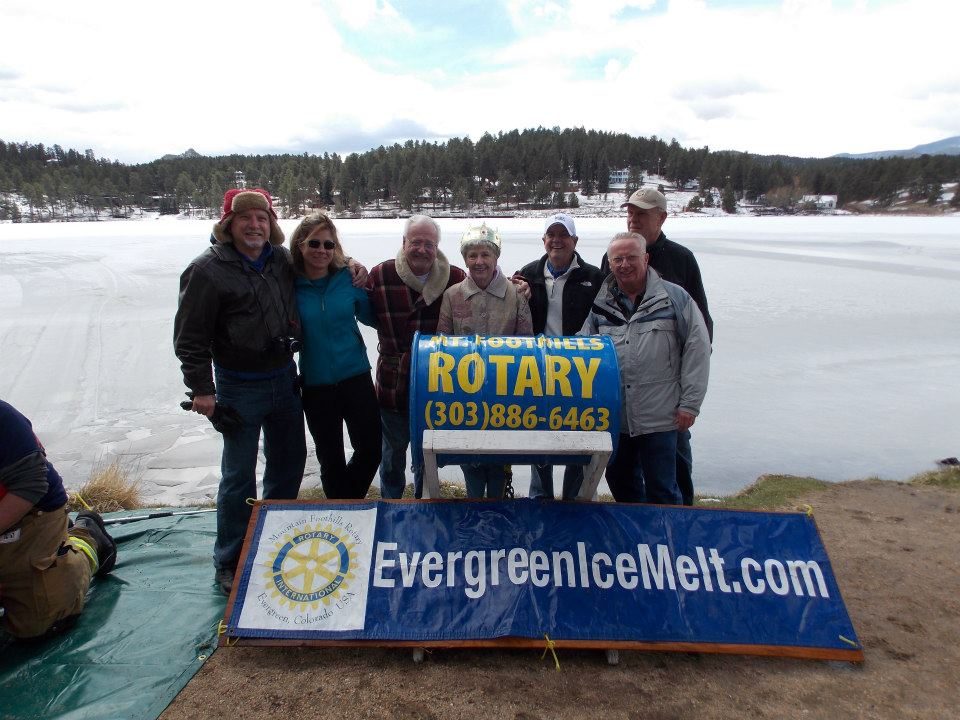Club members — including a freshly crowned Ice Melt winner — behind the Rotary barrel and EvergreenIceMelt banner on the shore of Evergreen Lake